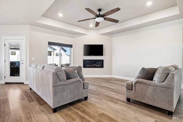 living room with a glass covered fireplace, light wood-type flooring, a raised ceiling, and baseboards