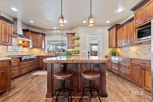 kitchen with wall chimney range hood, hanging light fixtures, stainless steel appliances, a center island, and light stone countertops