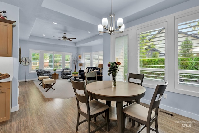 dining space with light hardwood / wood-style flooring, ceiling fan with notable chandelier, and a tray ceiling