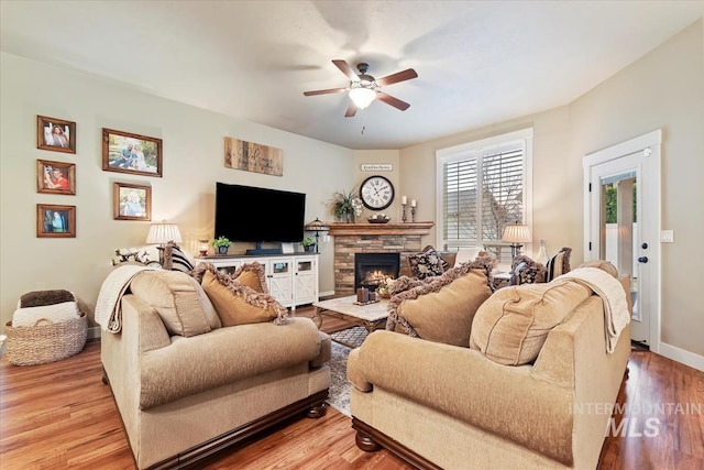 living area featuring ceiling fan, baseboards, a stone fireplace, and wood finished floors