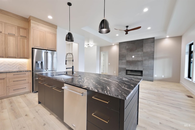 kitchen with stainless steel appliances, sink, light brown cabinets, a center island with sink, and hanging light fixtures