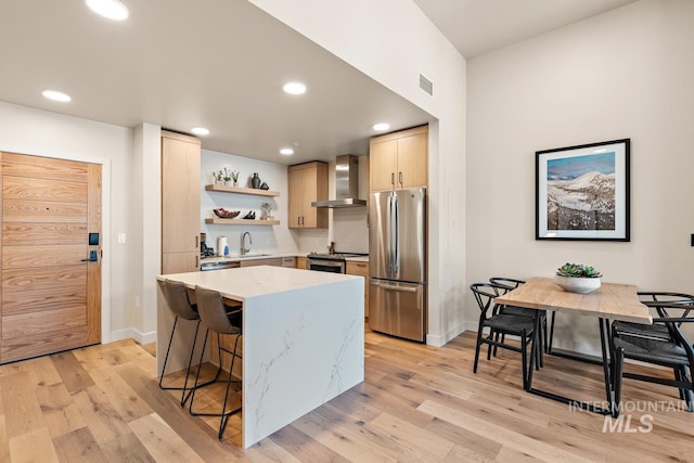 kitchen with wall chimney exhaust hood, stainless steel appliances, light brown cabinetry, and light wood-type flooring