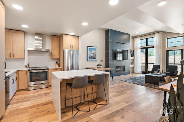 kitchen with light wood-type flooring, light brown cabinets, wall chimney exhaust hood, and appliances with stainless steel finishes