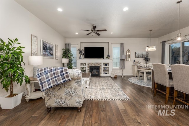 living room featuring a wealth of natural light, dark wood-style flooring, a glass covered fireplace, recessed lighting, and ceiling fan with notable chandelier