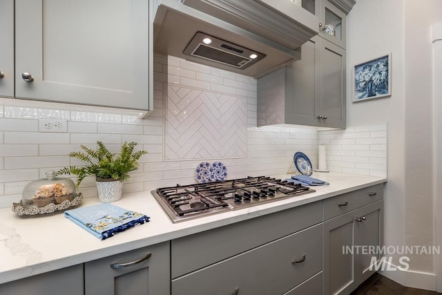 kitchen with stainless steel gas stovetop, gray cabinets, wall chimney exhaust hood, decorative backsplash, and glass insert cabinets