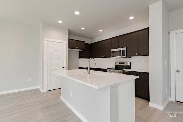 kitchen with appliances with stainless steel finishes, a kitchen island with sink, light wood-style flooring, and tasteful backsplash