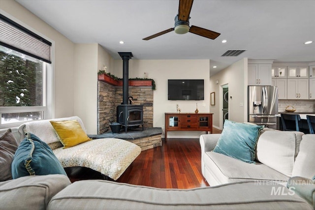 living room with dark hardwood / wood-style floors, a wood stove, ceiling fan, and stacked washer / drying machine