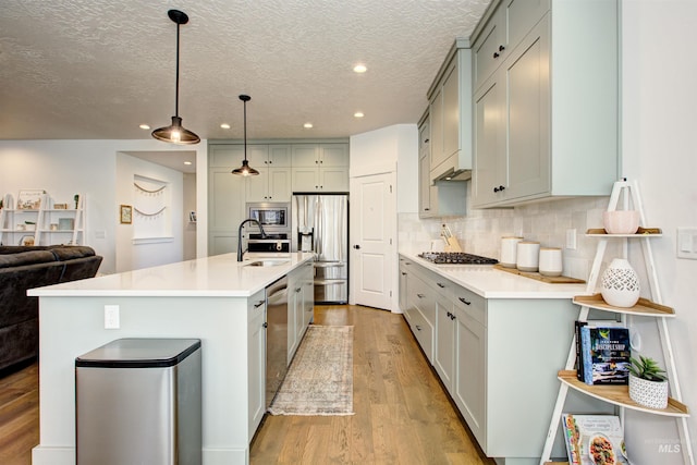 kitchen featuring appliances with stainless steel finishes, a kitchen island with sink, sink, decorative light fixtures, and light hardwood / wood-style flooring