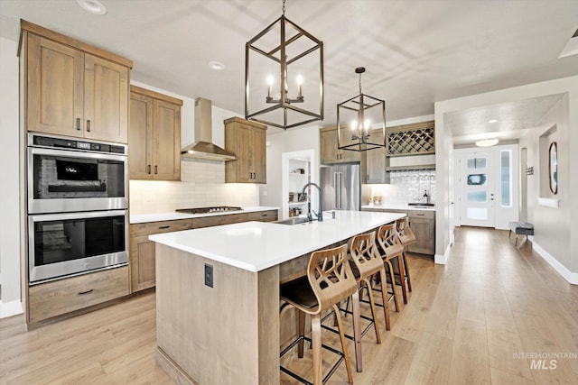 kitchen featuring light wood-style flooring, a sink, decorative backsplash, wall chimney range hood, and appliances with stainless steel finishes