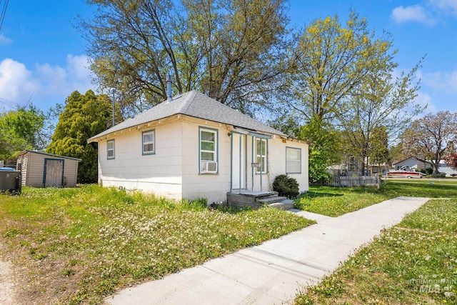 bungalow-style house featuring roof with shingles, a storage unit, a front lawn, cooling unit, and an outdoor structure