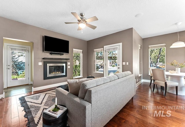 living area with dark wood finished floors, ceiling fan, baseboards, a glass covered fireplace, and a textured ceiling