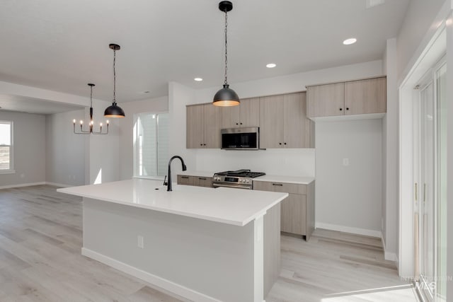 kitchen with stainless steel appliances, light brown cabinets, hanging light fixtures, an island with sink, and light hardwood / wood-style floors