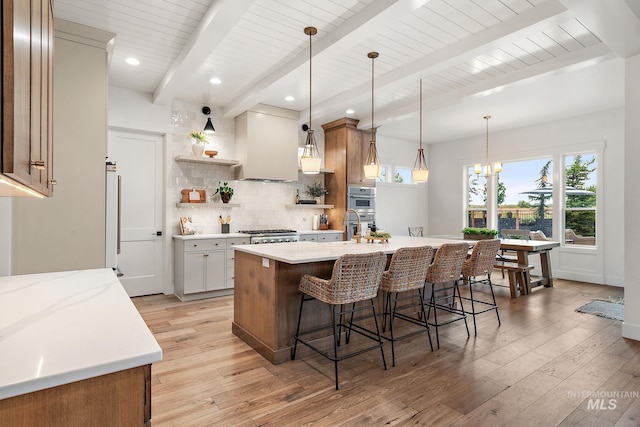 kitchen with decorative backsplash, light wood-type flooring, custom range hood, decorative light fixtures, and an island with sink