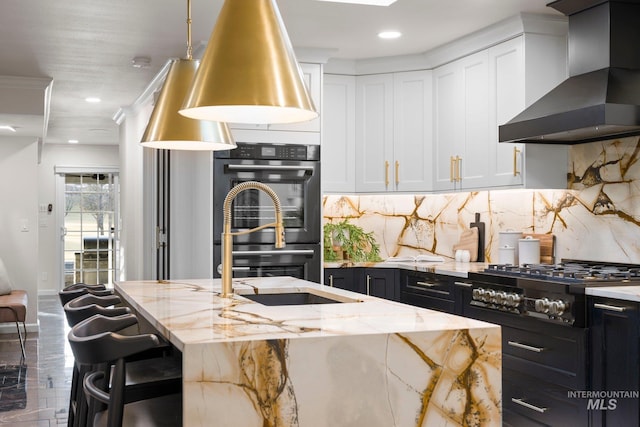 kitchen with white cabinets, wall chimney exhaust hood, dark cabinets, a kitchen island with sink, and black appliances