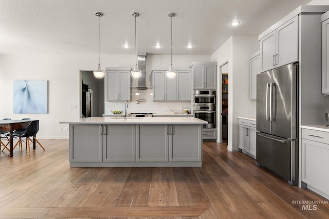 kitchen featuring stainless steel appliances, gray cabinets, an island with sink, and wall chimney range hood