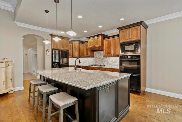 kitchen featuring a center island with sink, black appliances, ornamental molding, light stone counters, and light wood-type flooring