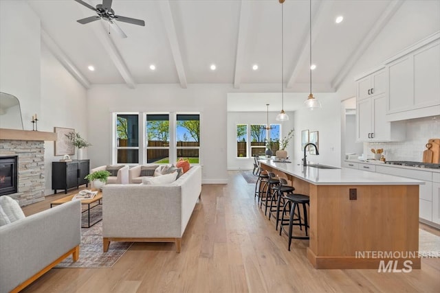 living room featuring ceiling fan, sink, a stone fireplace, light hardwood / wood-style flooring, and lofted ceiling with beams