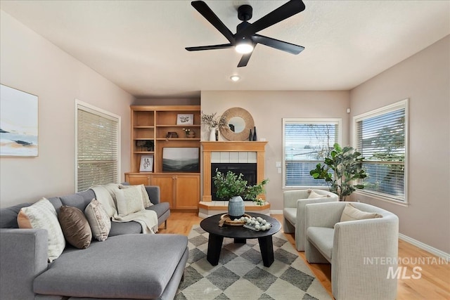 living area featuring light wood-style flooring, baseboards, ceiling fan, and a tiled fireplace