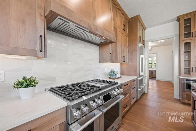 8860 Kemp Rd, Middleton, ID 83607 - kitchen featuring appliances with stainless steel finishes, custom exhaust hood, backsplash, glass insert cabinets, and light wood-style flooring