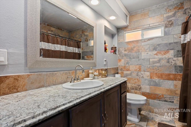 full bath featuring tile walls, stone tile flooring, a shower with shower curtain, toilet, and vanity