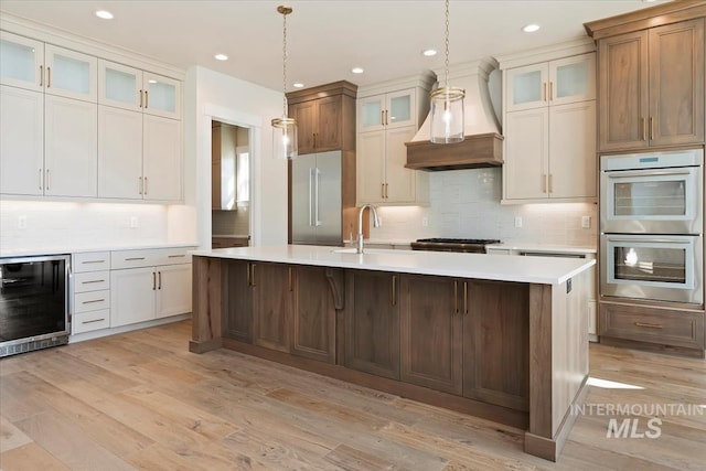 kitchen with wine cooler, custom exhaust hood, appliances with stainless steel finishes, pendant lighting, and a kitchen island with sink