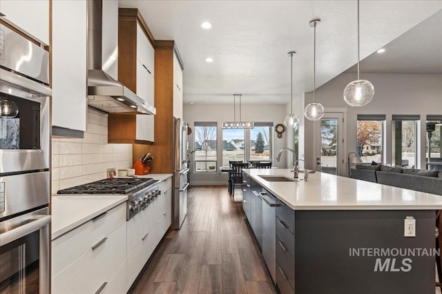 kitchen with decorative light fixtures, sink, white cabinets, a kitchen island with sink, and wall chimney range hood