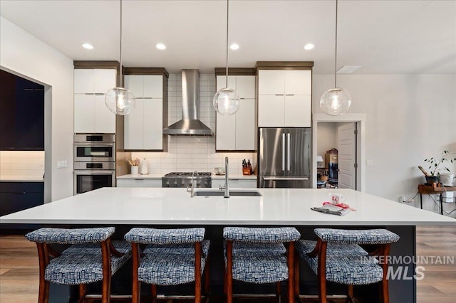 kitchen featuring stainless steel appliances, an island with sink, white cabinets, and wall chimney exhaust hood