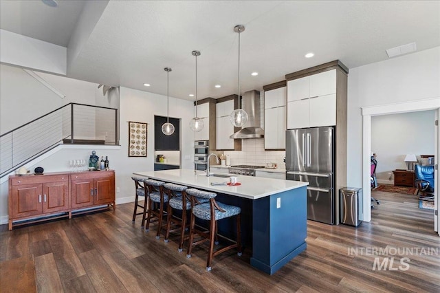 kitchen with appliances with stainless steel finishes, pendant lighting, white cabinetry, an island with sink, and wall chimney exhaust hood