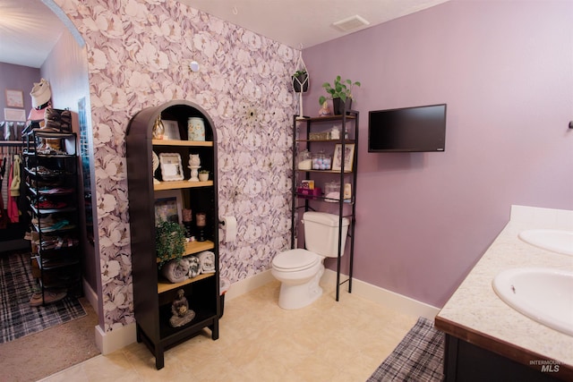 bathroom with double vanity, toilet, baseboards, wallpapered walls, and a walk in closet