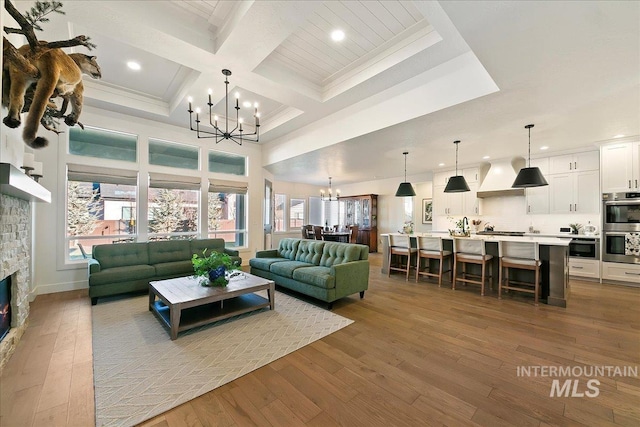 living room with an inviting chandelier, wood-type flooring, coffered ceiling, and a stone fireplace