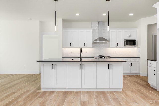 kitchen with an island with sink, stainless steel microwave, hanging light fixtures, and wall chimney exhaust hood