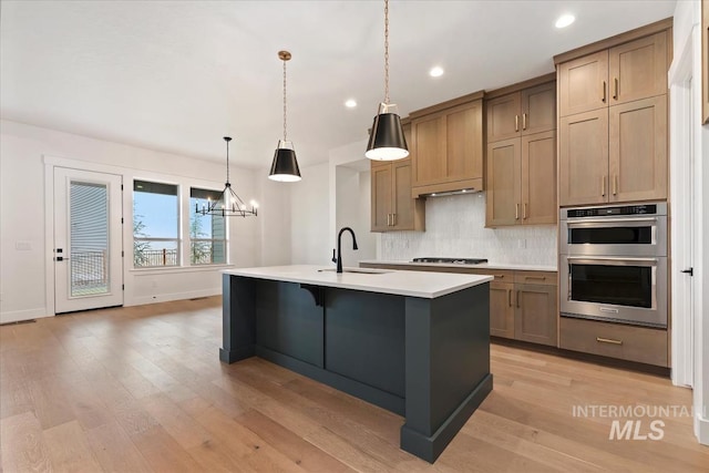 kitchen featuring an island with sink, light wood-style flooring, a sink, stainless steel double oven, and backsplash