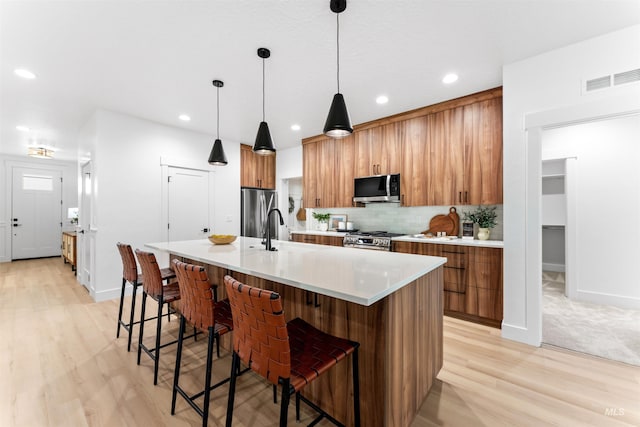 kitchen featuring sink, stainless steel appliances, decorative light fixtures, a center island with sink, and light wood-type flooring