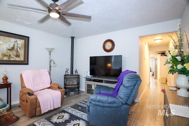 living room featuring a wood stove, ceiling fan, visible vents, wood finished floors, and baseboards