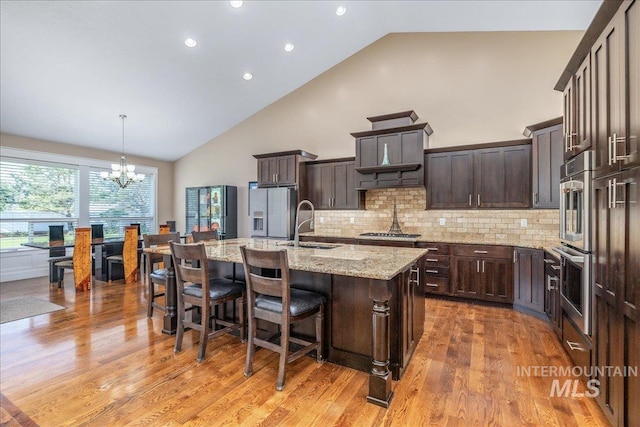 kitchen with dark brown cabinetry, an inviting chandelier, high vaulted ceiling, an island with sink, and stainless steel appliances