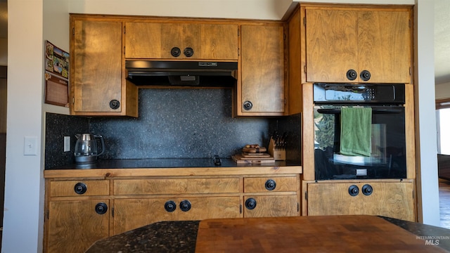 kitchen featuring under cabinet range hood, black oven, brown cabinets, tasteful backsplash, and dark countertops