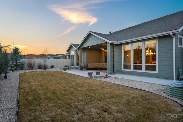 back of house at dusk with a patio, a lawn, roof with shingles, and fence