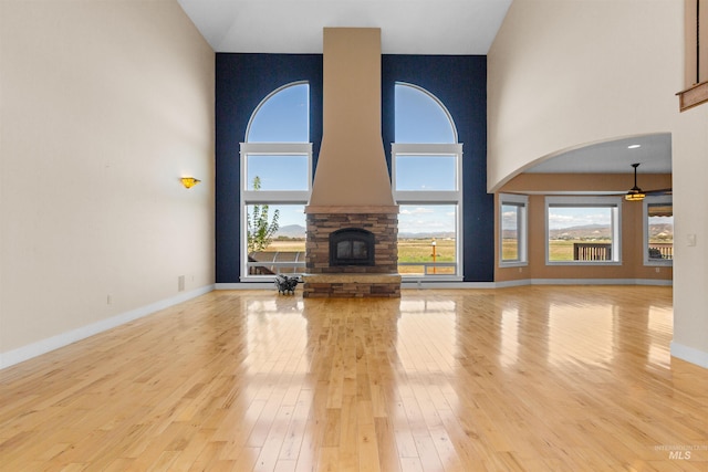 unfurnished living room with ceiling fan, a stone fireplace, a towering ceiling, baseboards, and wood-type flooring