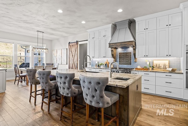 kitchen with an island with sink, decorative backsplash, light wood-style floors, custom exhaust hood, and white cabinetry