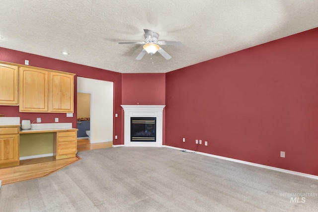 unfurnished living room featuring a ceiling fan, light colored carpet, a textured ceiling, a glass covered fireplace, and baseboards