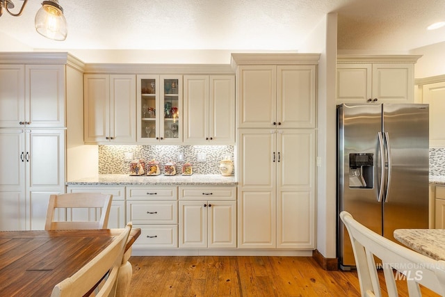 kitchen with stainless steel refrigerator with ice dispenser, light wood-type flooring, light stone countertops, cream cabinets, and backsplash