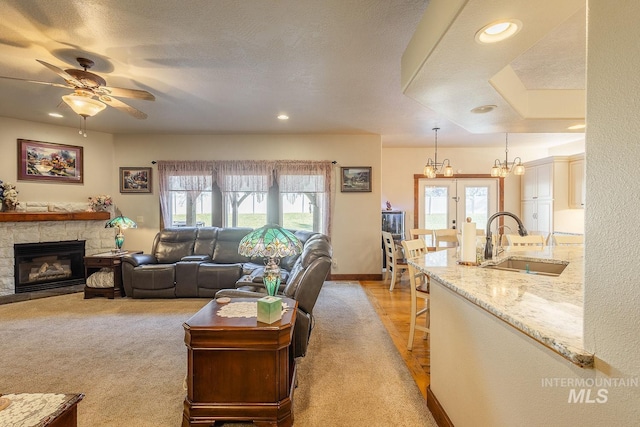 carpeted living room featuring a stone fireplace, sink, ceiling fan with notable chandelier, and a textured ceiling