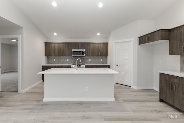 kitchen featuring tasteful backsplash, dark brown cabinetry, a kitchen island with sink, stainless steel microwave, and light countertops