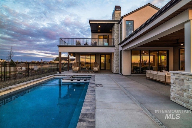 back of house at dusk featuring a chimney, fence, a balcony, a patio, and stone siding