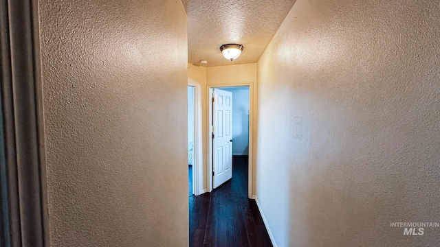 hallway featuring a textured ceiling, a textured wall, baseboards, and dark wood-style floors
