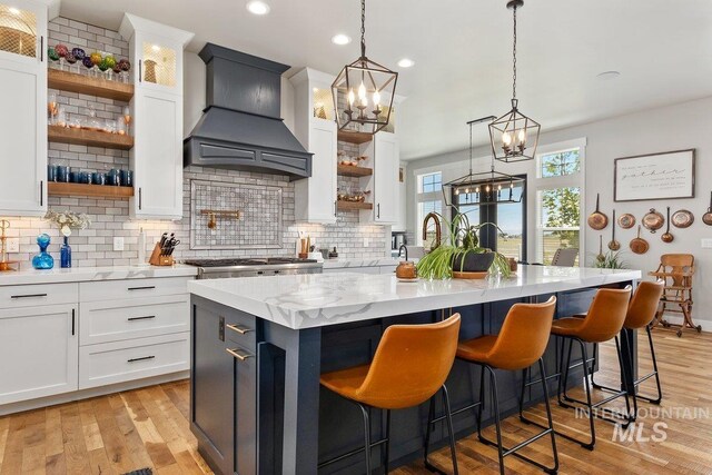 kitchen with pendant lighting, custom exhaust hood, white cabinets, a center island with sink, and light hardwood / wood-style flooring