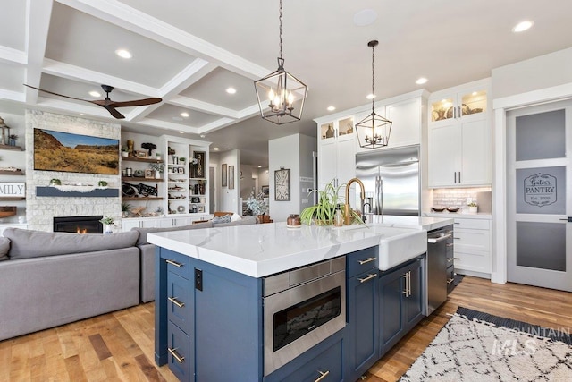 kitchen featuring coffered ceiling, a center island with sink, a stone fireplace, built in appliances, and white cabinetry