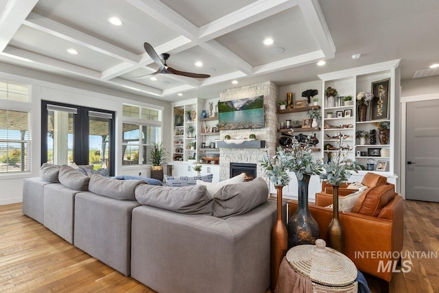 living room featuring beam ceiling, ceiling fan, coffered ceiling, a stone fireplace, and light hardwood / wood-style flooring