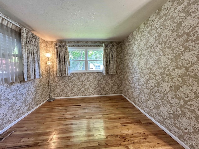 spare room with a textured ceiling, wallpapered walls, baseboards, visible vents, and hardwood / wood-style flooring