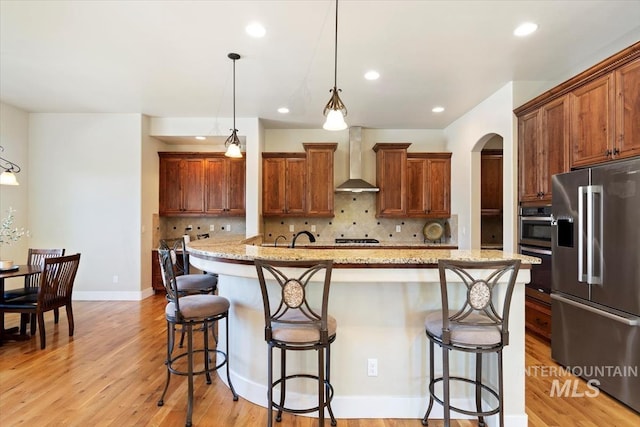 kitchen with stainless steel appliances, wall chimney range hood, light wood-type flooring, decorative backsplash, and an island with sink